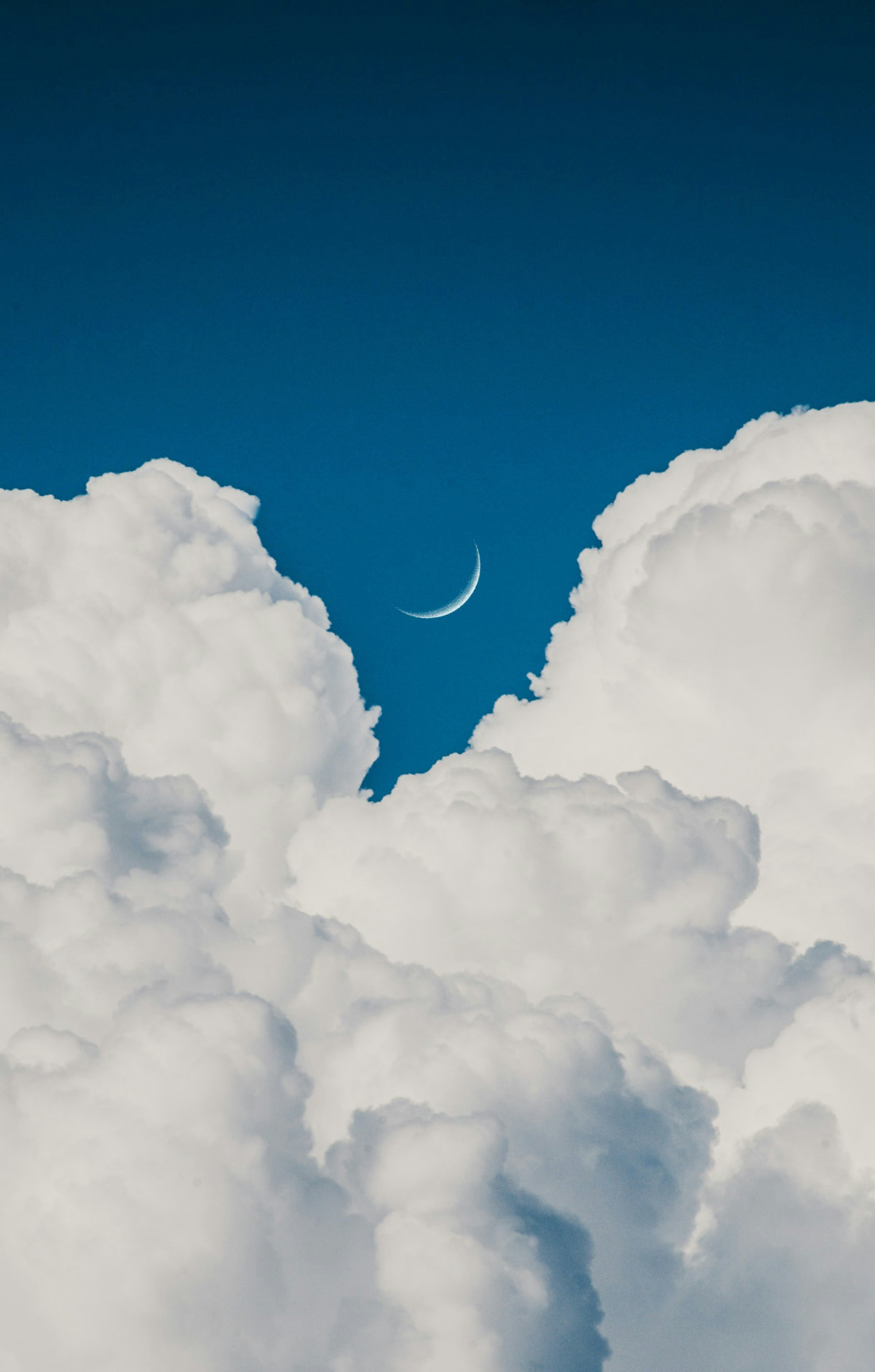 A slender crescent moon framed by billowing clouds in a deep blue sky